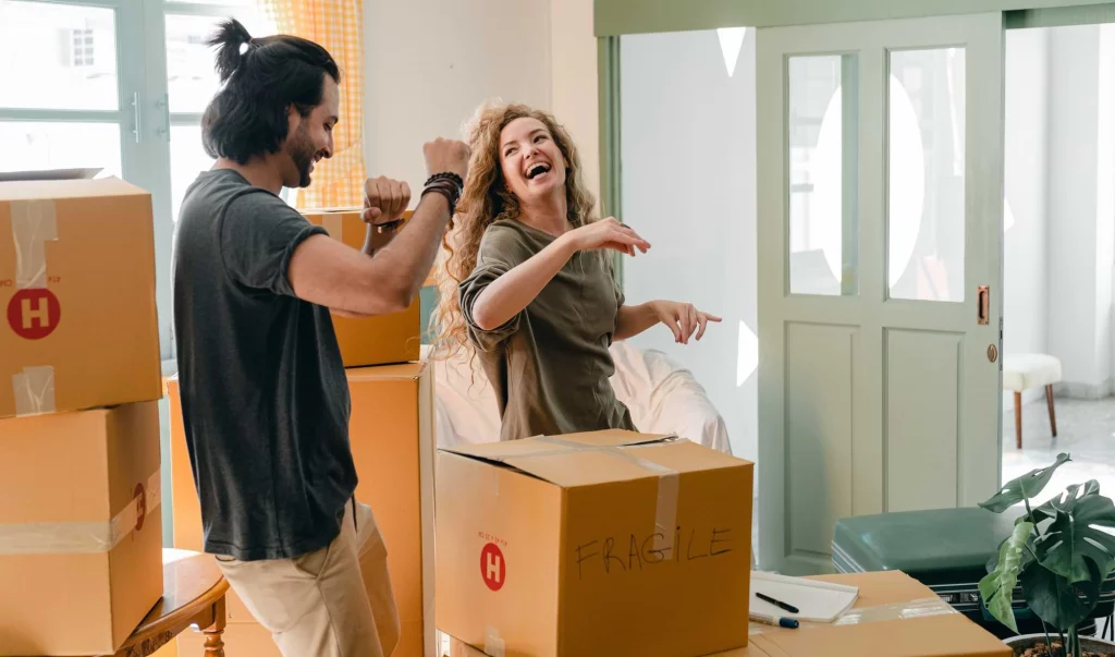 A couple is standing in the middle of an empty room, laughing, while surrounded by packed boxes.